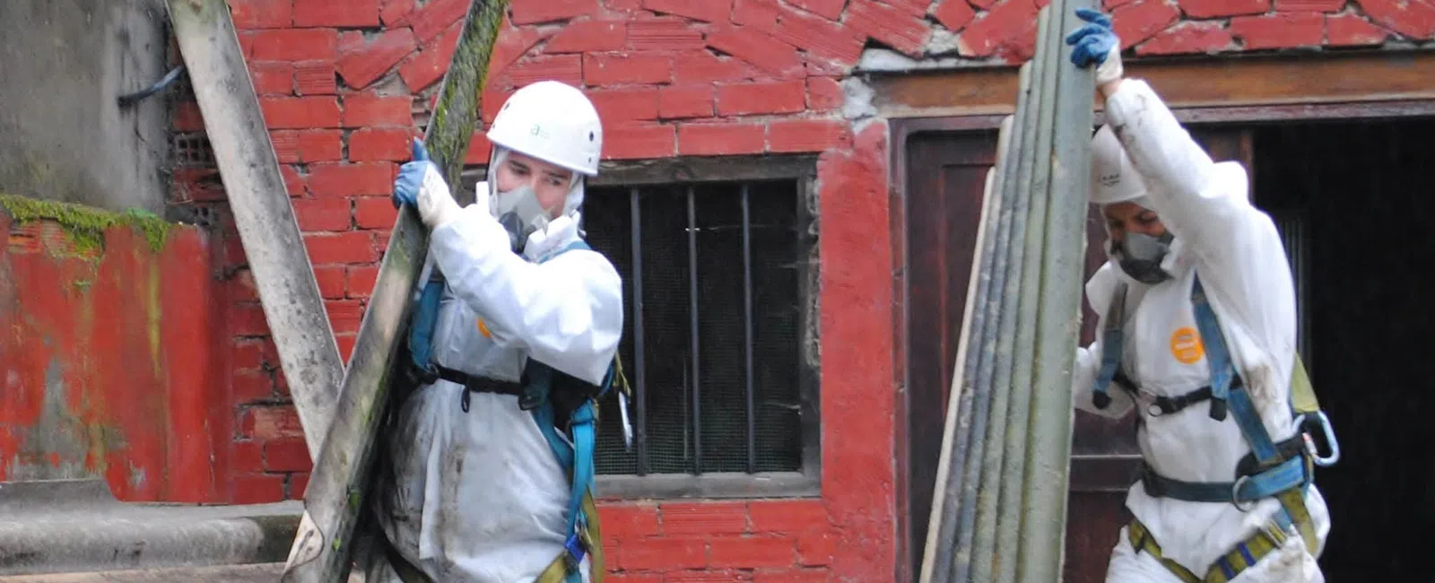 Two workers in protective suits and helmets removing corrugated asbestos sheets from a site with red brick walls.