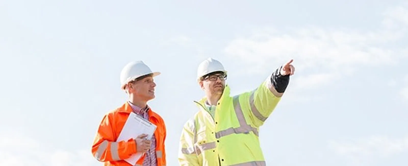 Two construction workers in safety jackets and helmets discussing at a construction site with one pointing ahead.