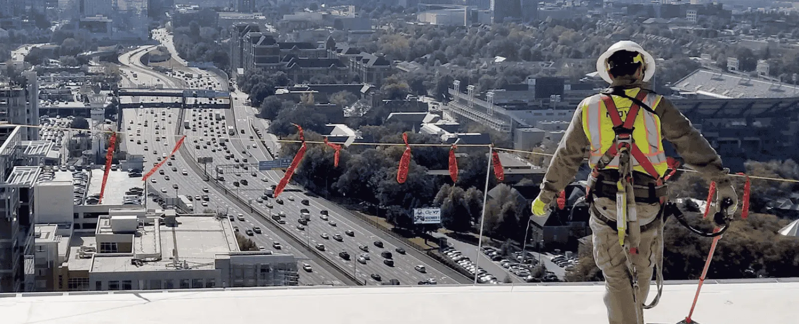 Construction worker in safety harness overlooking city skyline and busy highway from rooftop edge on clear day