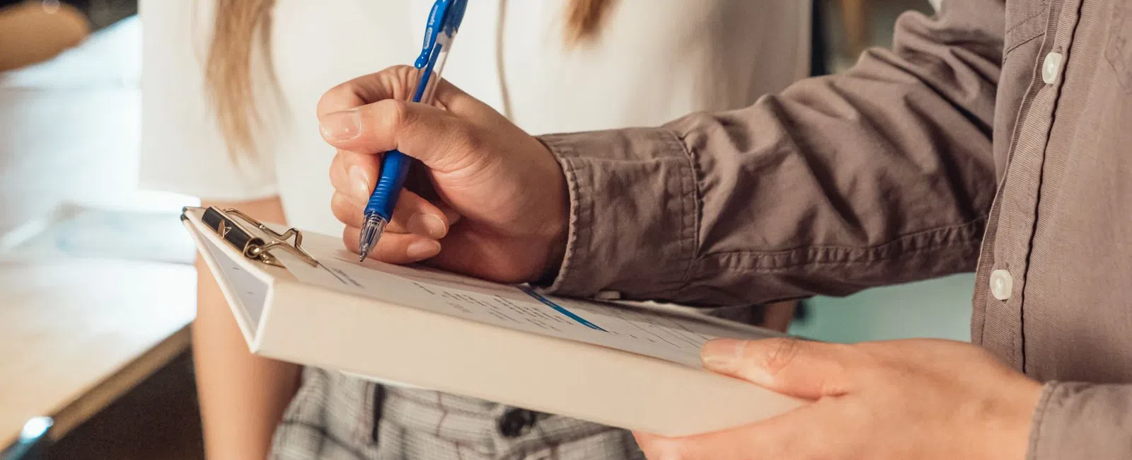 Person holding clipboard and pen while another stands close by, engaged in discussion or signing document.