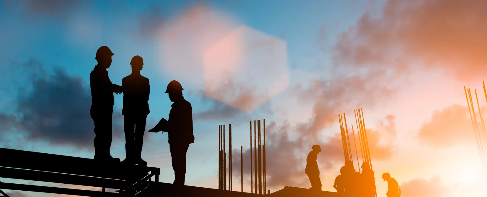 Silhouettes of construction workers on scaffolding during a sunset sky with clouds and sunlight flare.