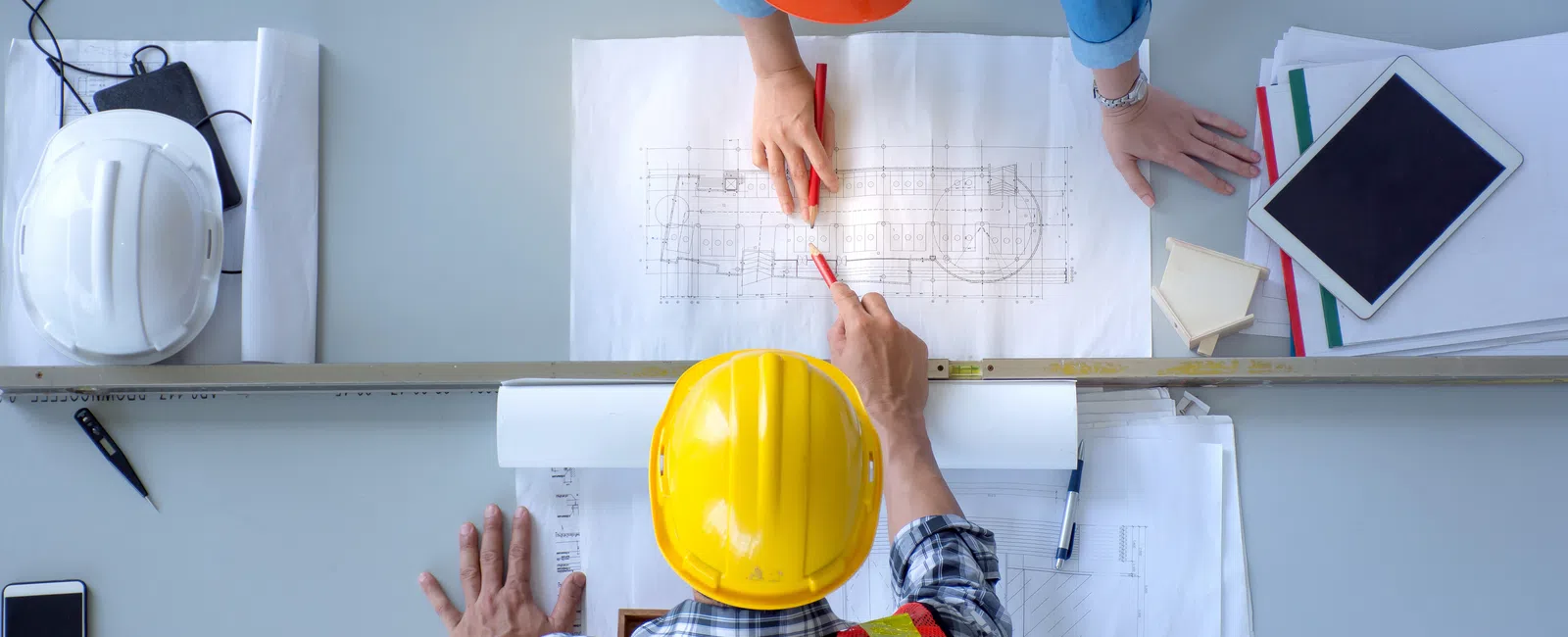 Construction workers in helmets and vests reviewing architectural blueprints on a table with tools and devices