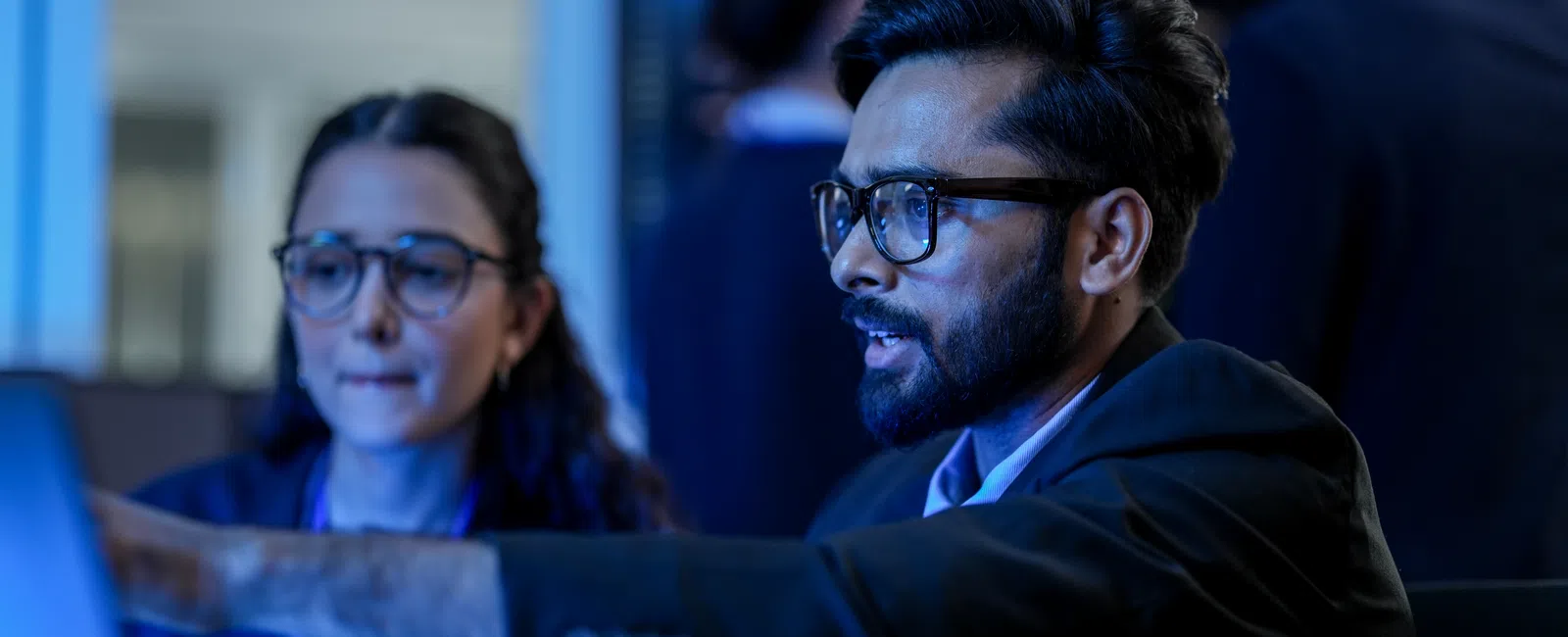 Two professionals wearing glasses collaborating and pointing at a computer screen in an office setting at night