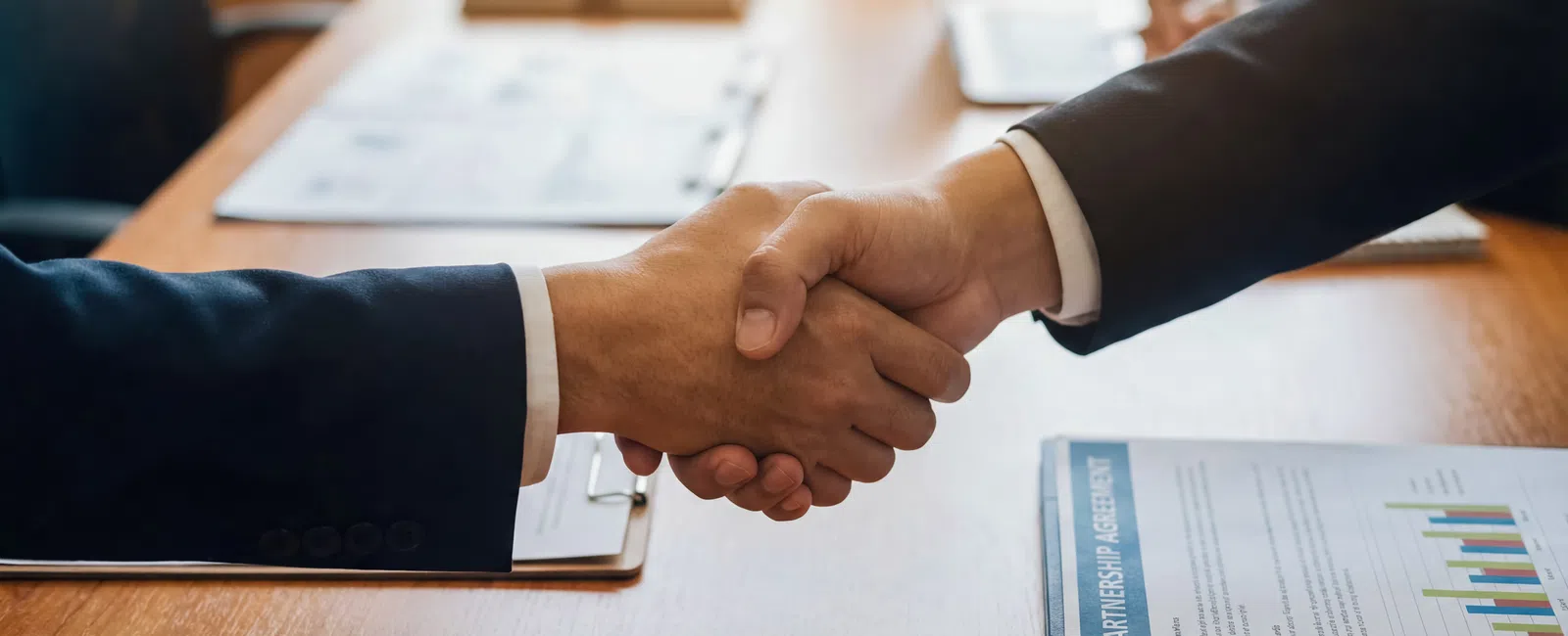 Two business professionals shaking hands over a wooden table with contract documents and charts.