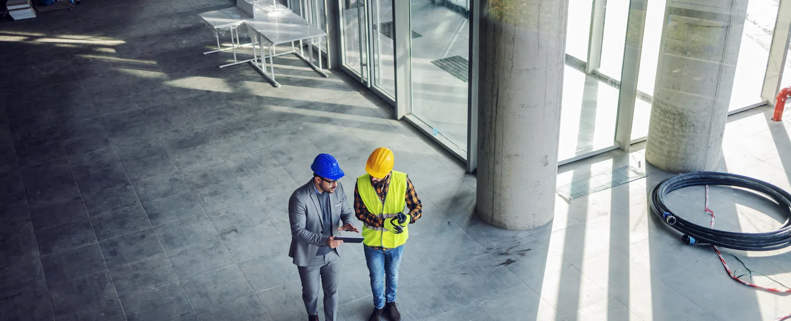 Two construction workers in helmets discuss plans inside a large modern building with glass walls and concrete columns.