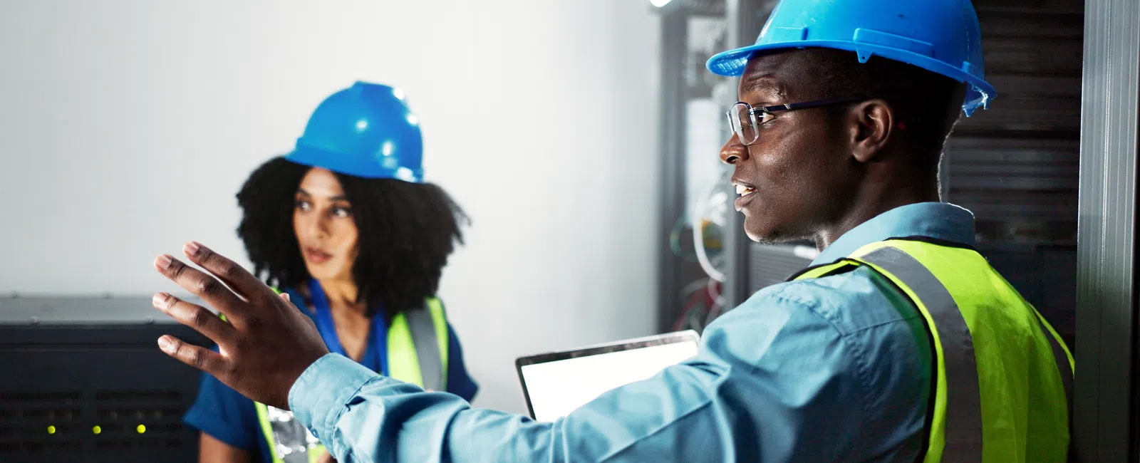 Two engineers in blue helmets and safety vests discuss technical details inside an industrial control room.