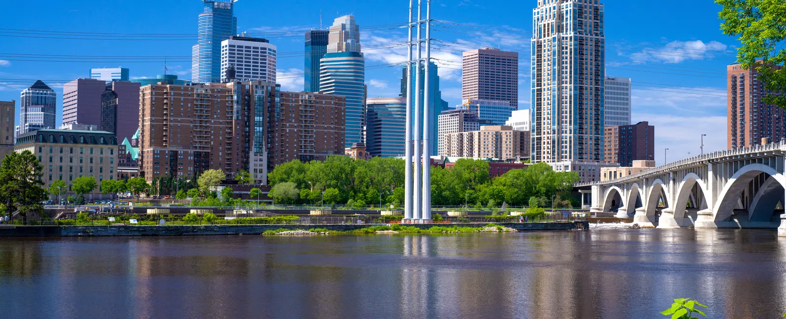 Minneapolis skyline with Stone Arch Bridge and Mississippi River under a clear blue sky with green trees.