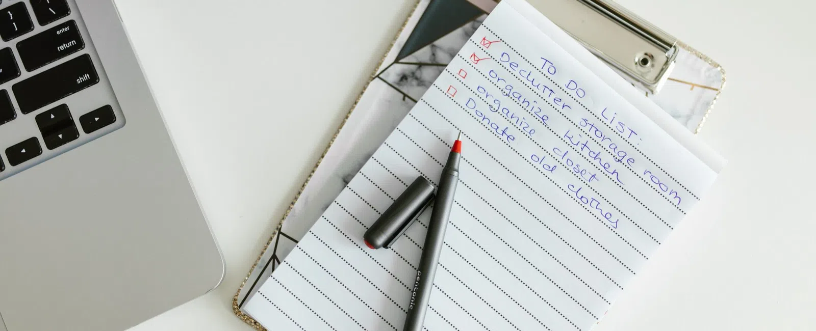 Clipboard with handwritten to-do list and pen next to laptop on a white desk workspace.