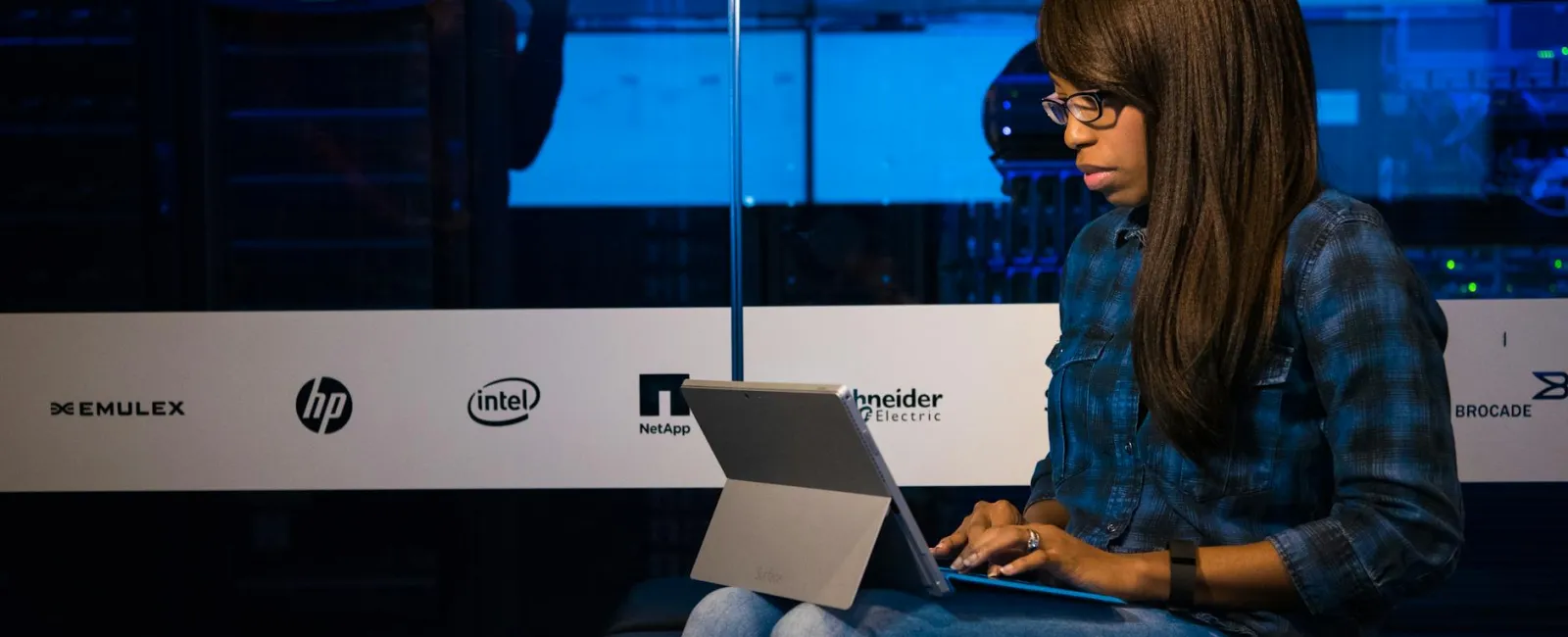 Young woman wearing glasses working on a laptop in a server room with technology company logos on the glass panel.