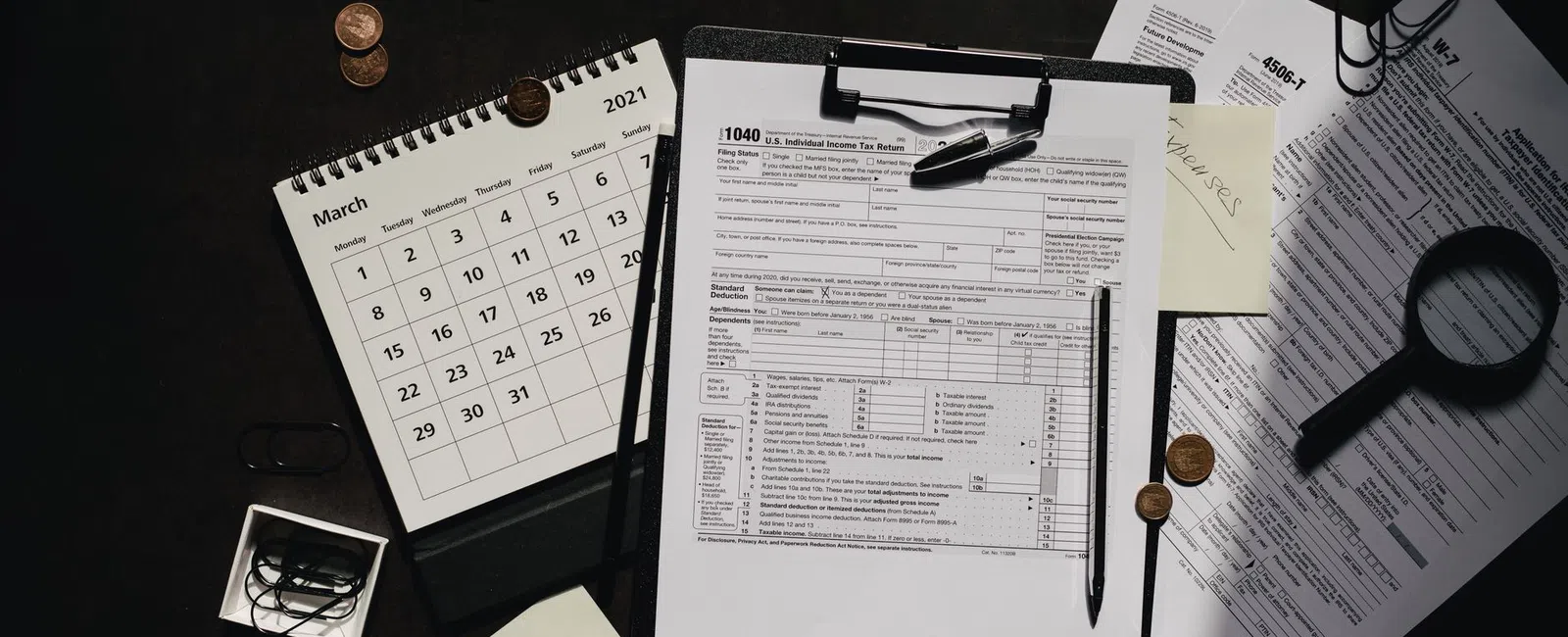 U.S. tax documents with pen, calendar showing March 2021, coins, paper clips, and magnifying glass on black surface