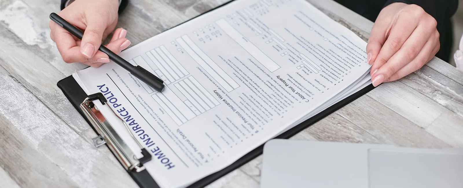 Person holding pen and pointing at home insurance policy form on clipboard with laptop on wooden table.