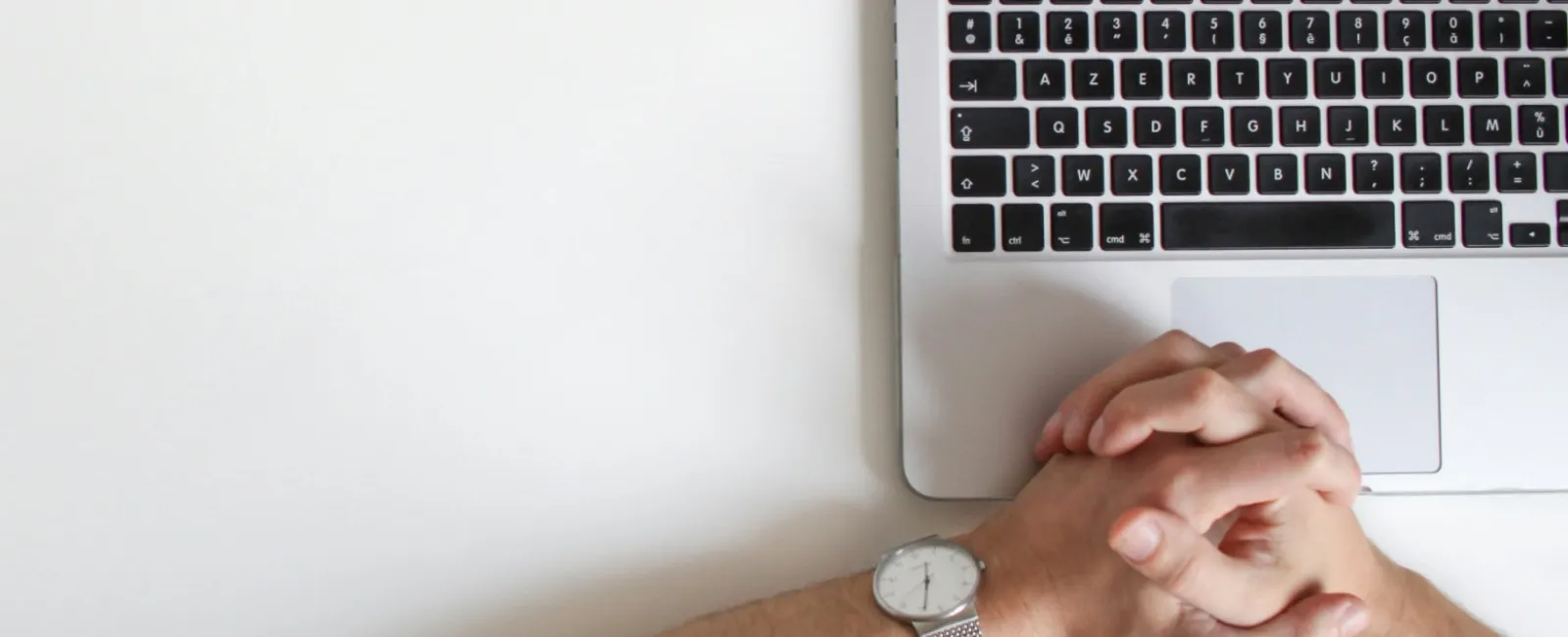 Person with folded hands resting beside a laptop keyboard on a white desk, wearing a silver watch.