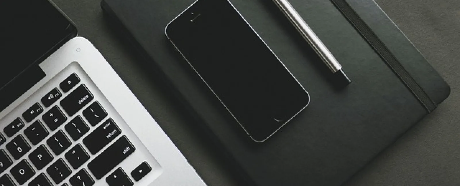 Laptop keyboard, smartphone on closed black notebook, and silver pen arranged on dark surface