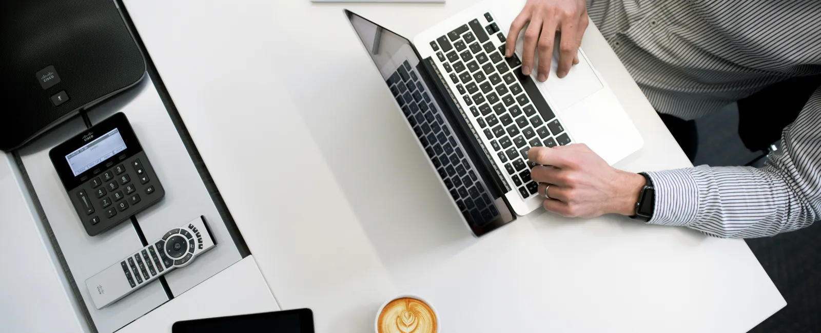 Person working on laptop at white desk with coffee, tablet, phone, remote, and notepad with pens.