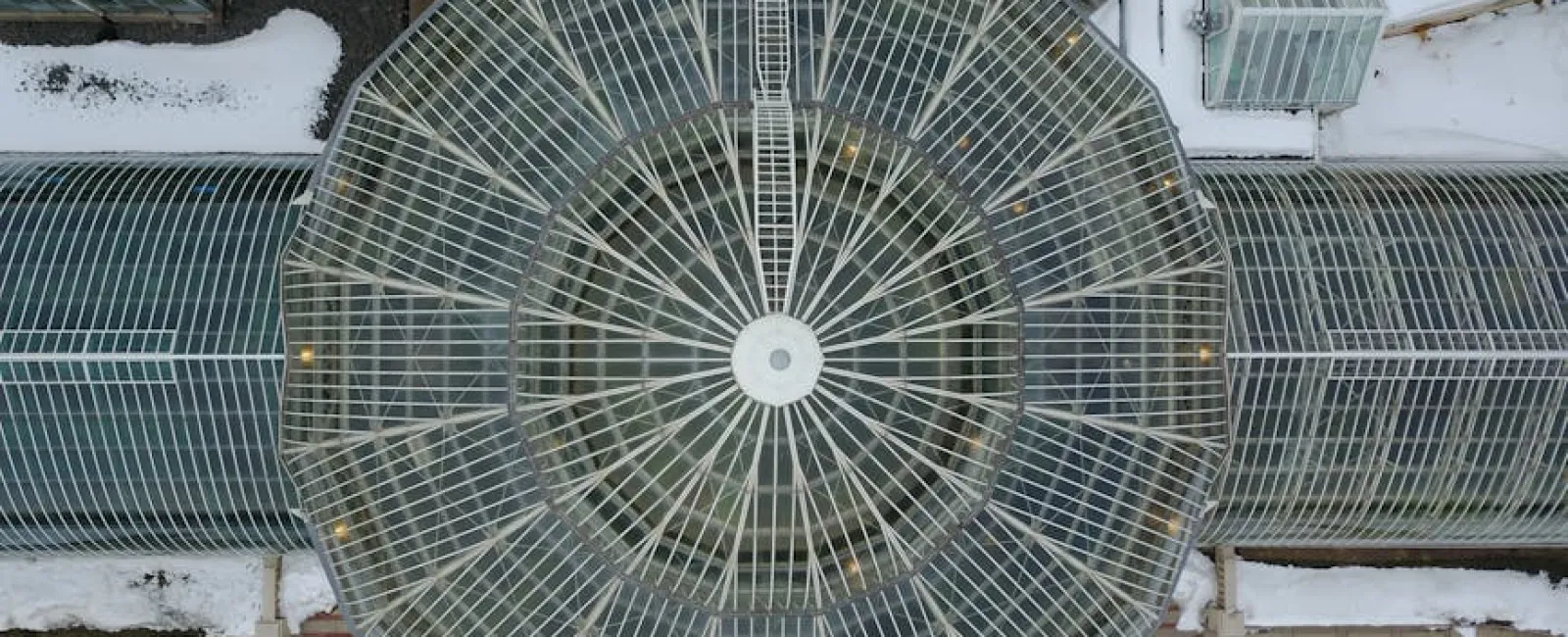 Aerial view of a large circular glass greenhouse structure with surrounding glass tunnels and snow-covered ground.