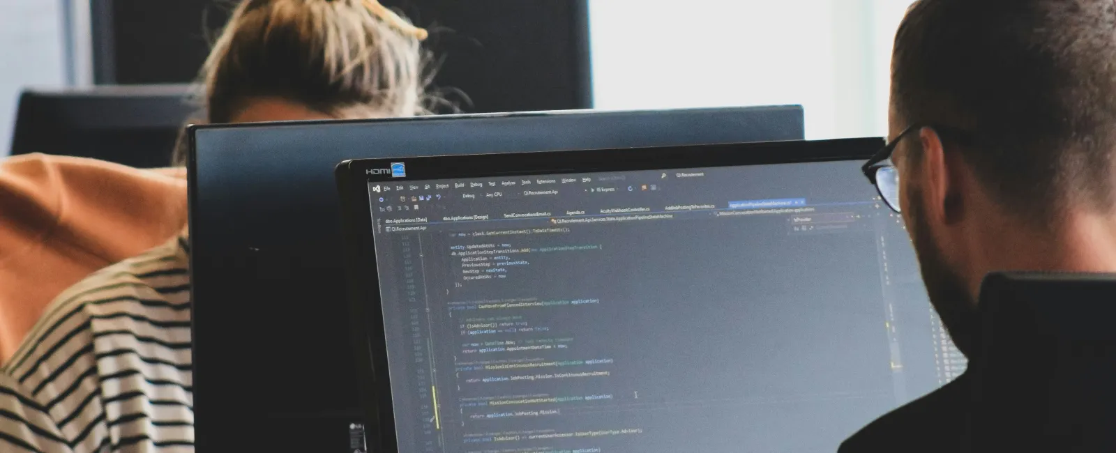 Two programmers working on computers with one screen displaying code in a software development environment.