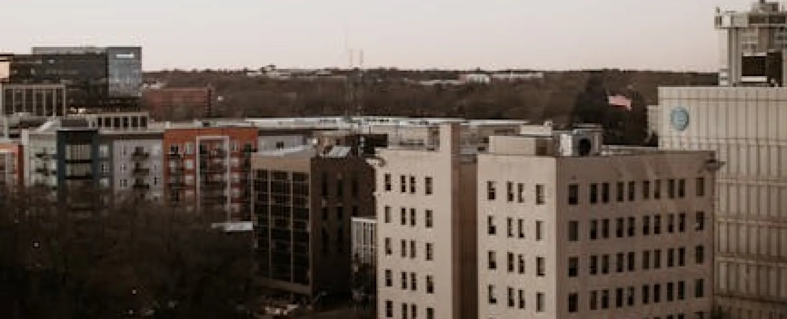 Urban cityscape featuring mid-rise buildings and rooftops under a pale sky during early evening.