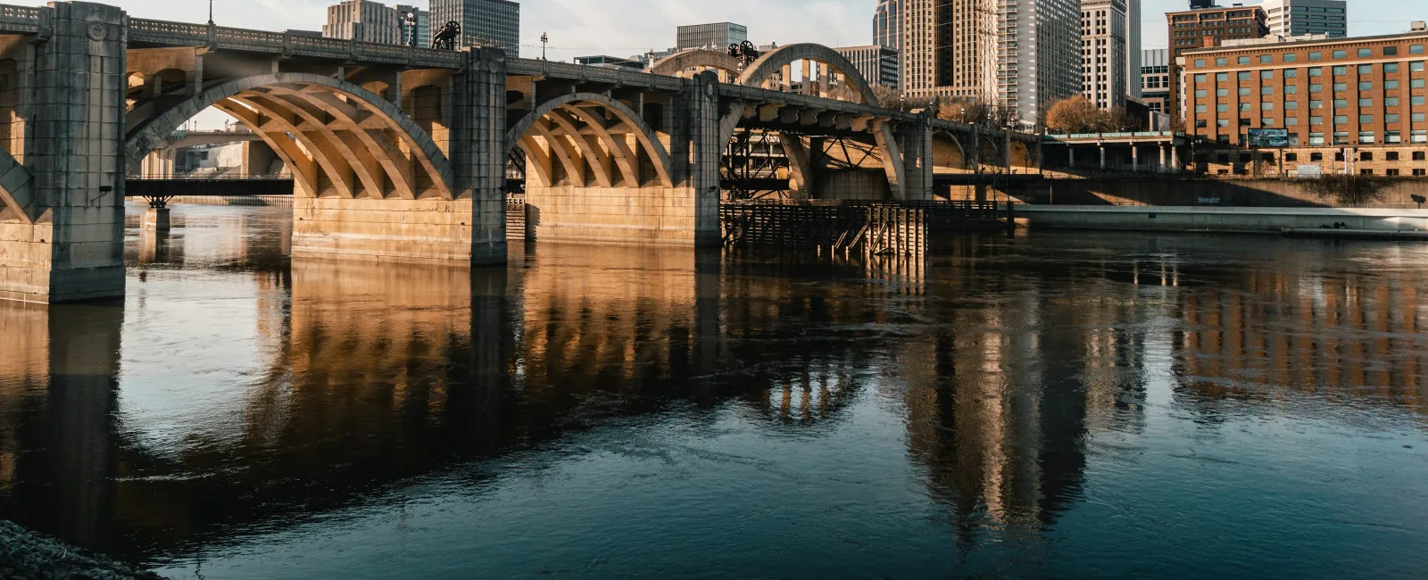 Historic stone arch bridge over calm river with city skyline and buildings in warm daylight reflection.