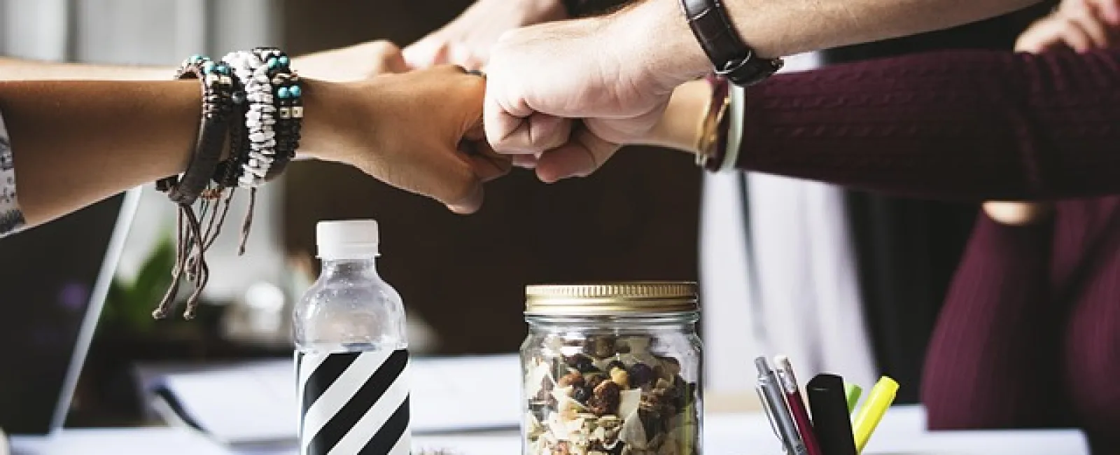 Diverse team of professionals joining fists over a work desk with snacks and office supplies