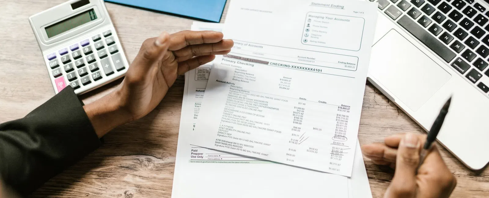 Person reviewing financial documents with calculator and laptop on wooden desk workspace.