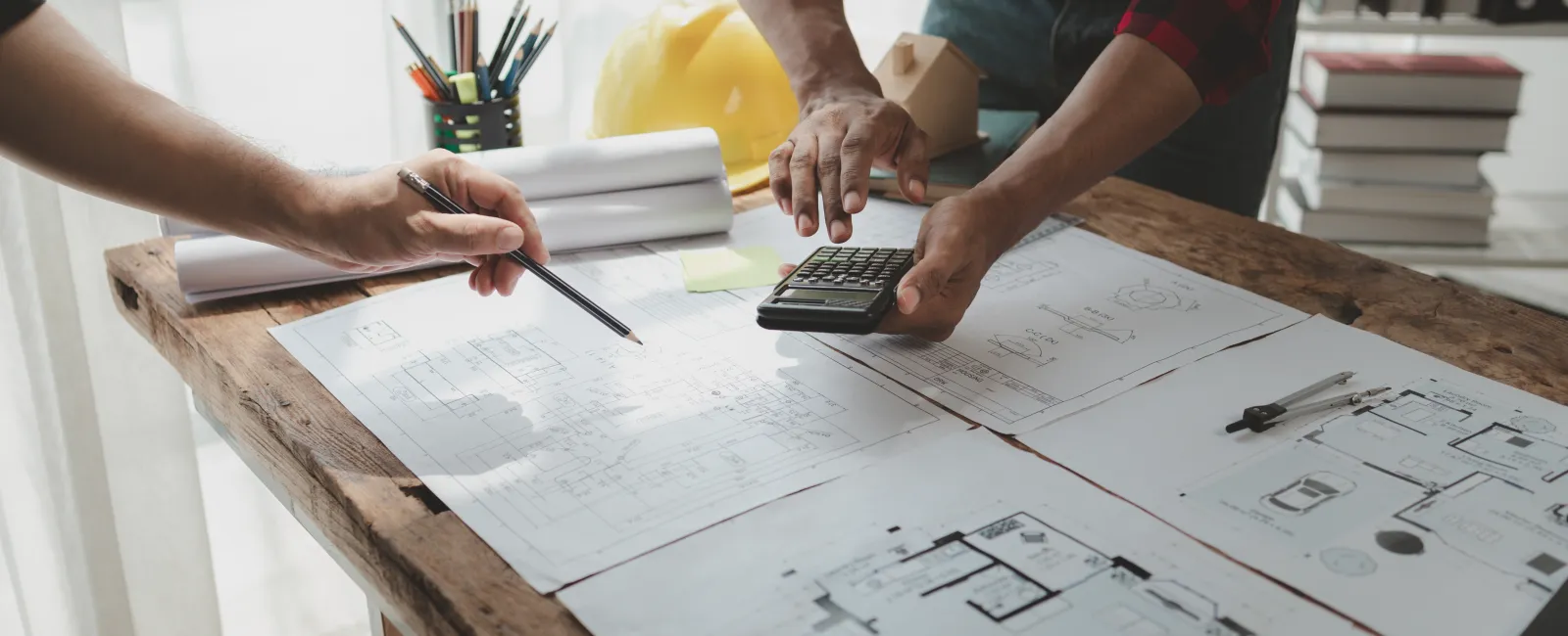 Two architects discussing building plans with a calculator, blueprints, and design tools on a wooden table.