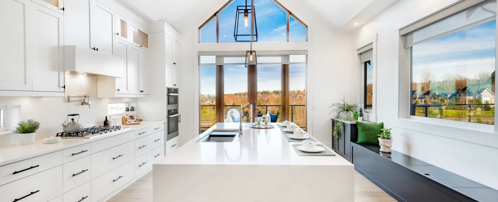 Bright modern kitchen with large white island, vaulted windows, black fixtures, and scenic outdoor views.
