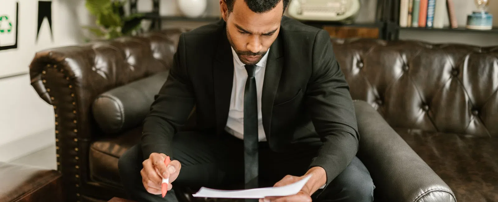 Man in black suit and tie sitting on leather sofa reviewing documents at wooden coffee table in office.