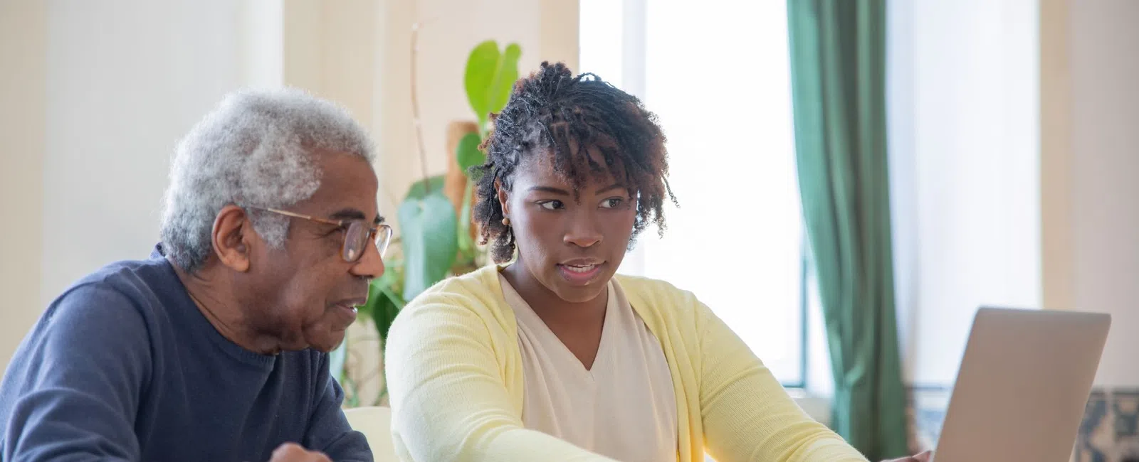 Young woman helping elderly man with laptop at home in a bright room with plants and sunlight.