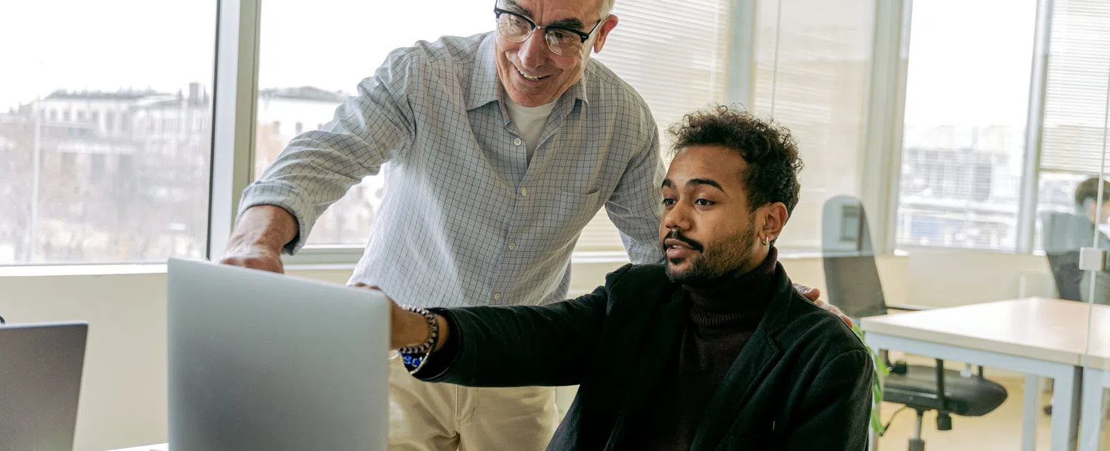 Two male colleagues collaborate over a laptop in a bright modern office with large windows during daytime.