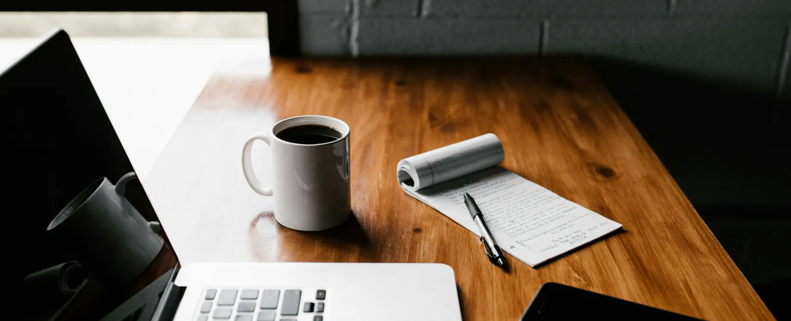 Laptop, coffee mug, notepad with pen, and smartphone on wooden desk in a cozy workspace near window.