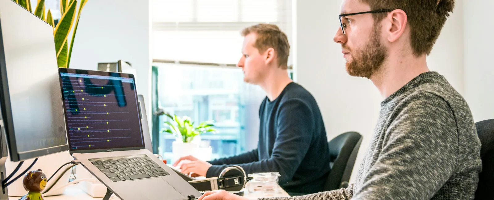 Two men working at computers in a bright modern office with plants and tech accessories on desks.