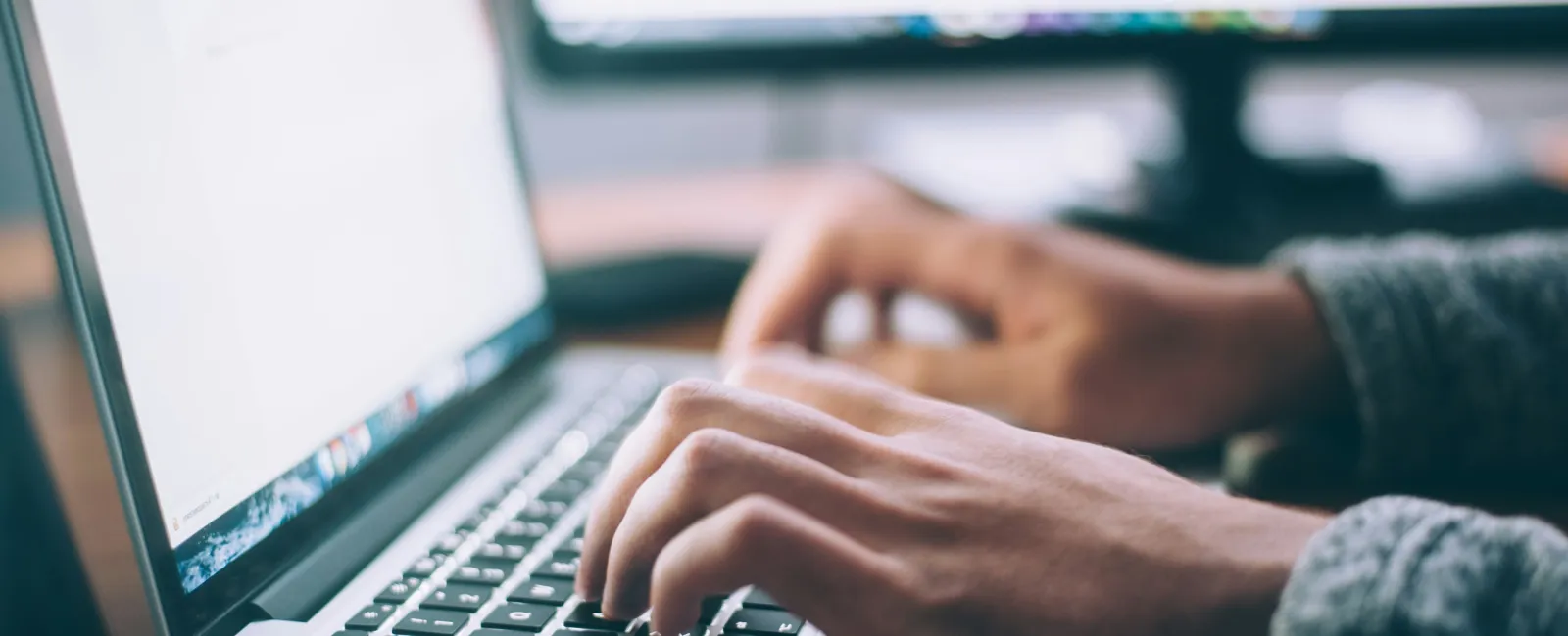 Hands typing on a laptop keyboard with a blurred monitor in the background on a wooden desk.