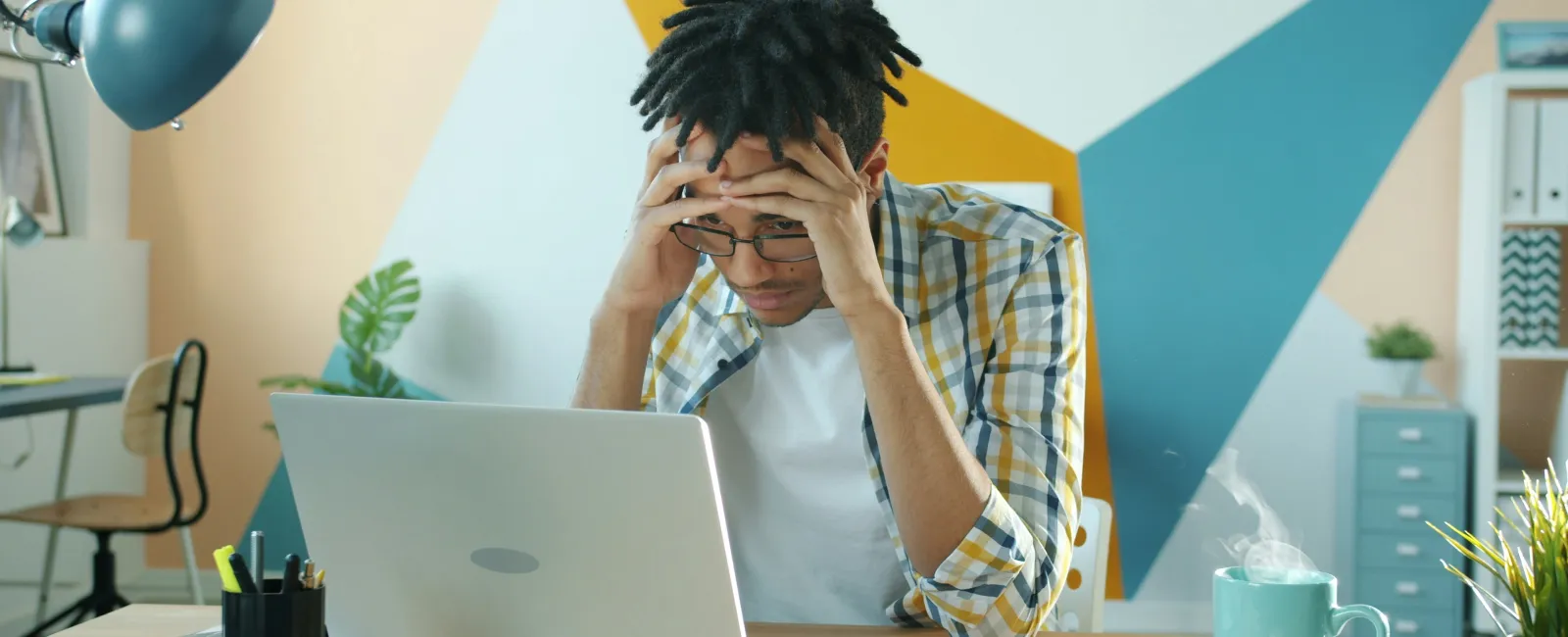 Stressed young man wearing glasses sitting at desk with laptop and coffee cup in colorful modern office.