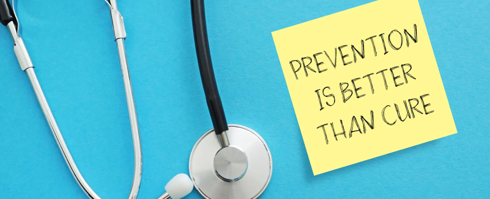 Stethoscope beside sticky note reading Prevention is better than cure on blue background.