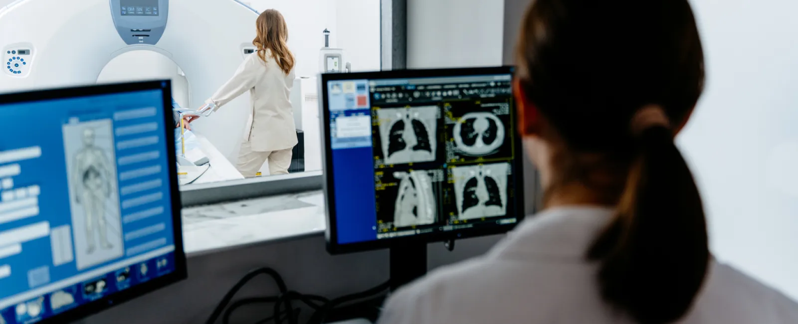Radiology technician analyzing lung CT scans on computer screens as patient prepares for MRI scan.