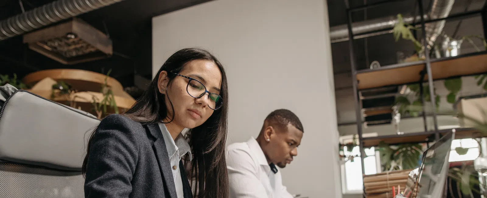 Two professionals working at a shared office desk with laptops, calculator, and documents in a modern workspace.