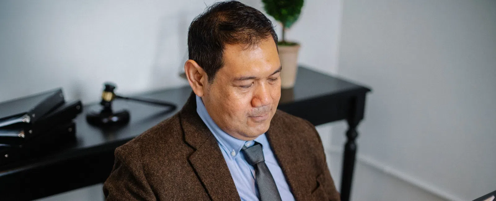 Man in business attire reviewing documents in a folder at office desk with files and plant in background.