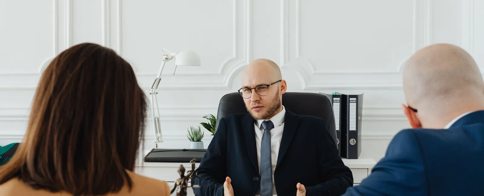Bald lawyer in suit consulting with a couple in formal attire in a bright office with legal scales on desk.