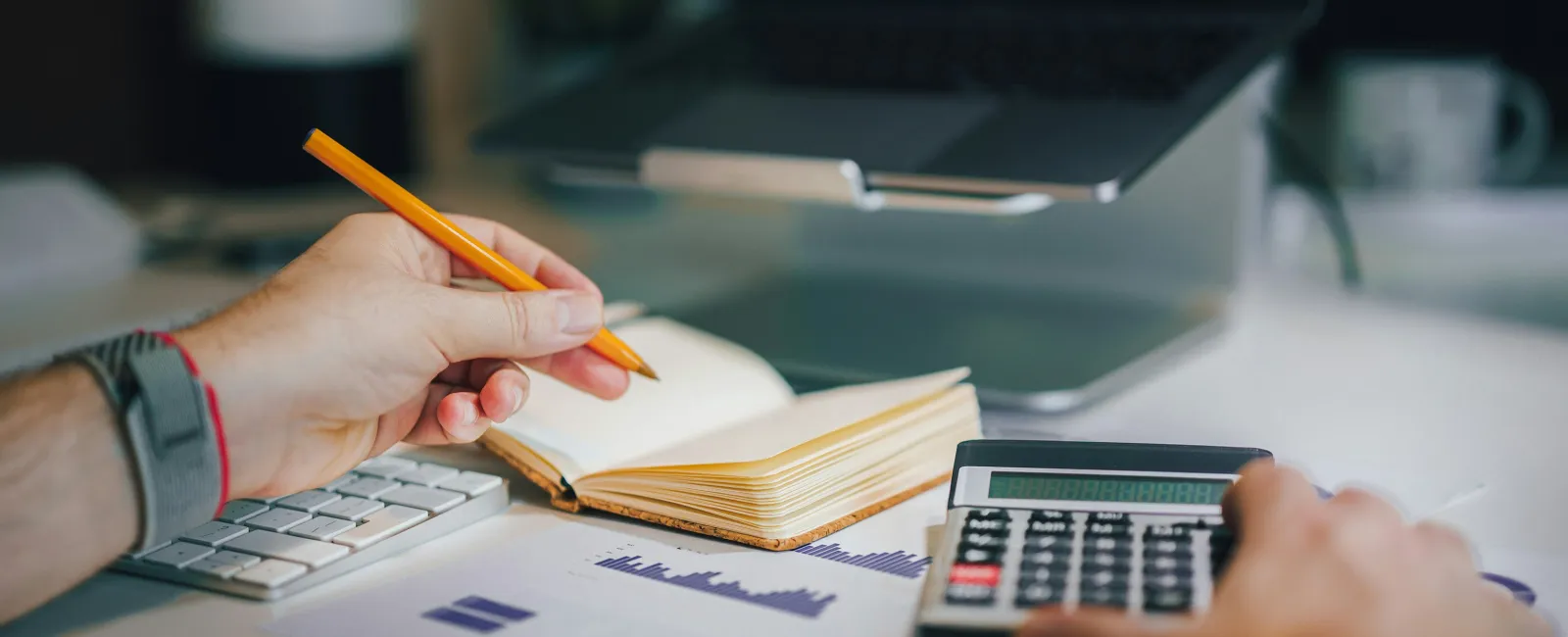 Person using calculator and pencil to analyze financial charts and notes at a desk with computer and keyboard.