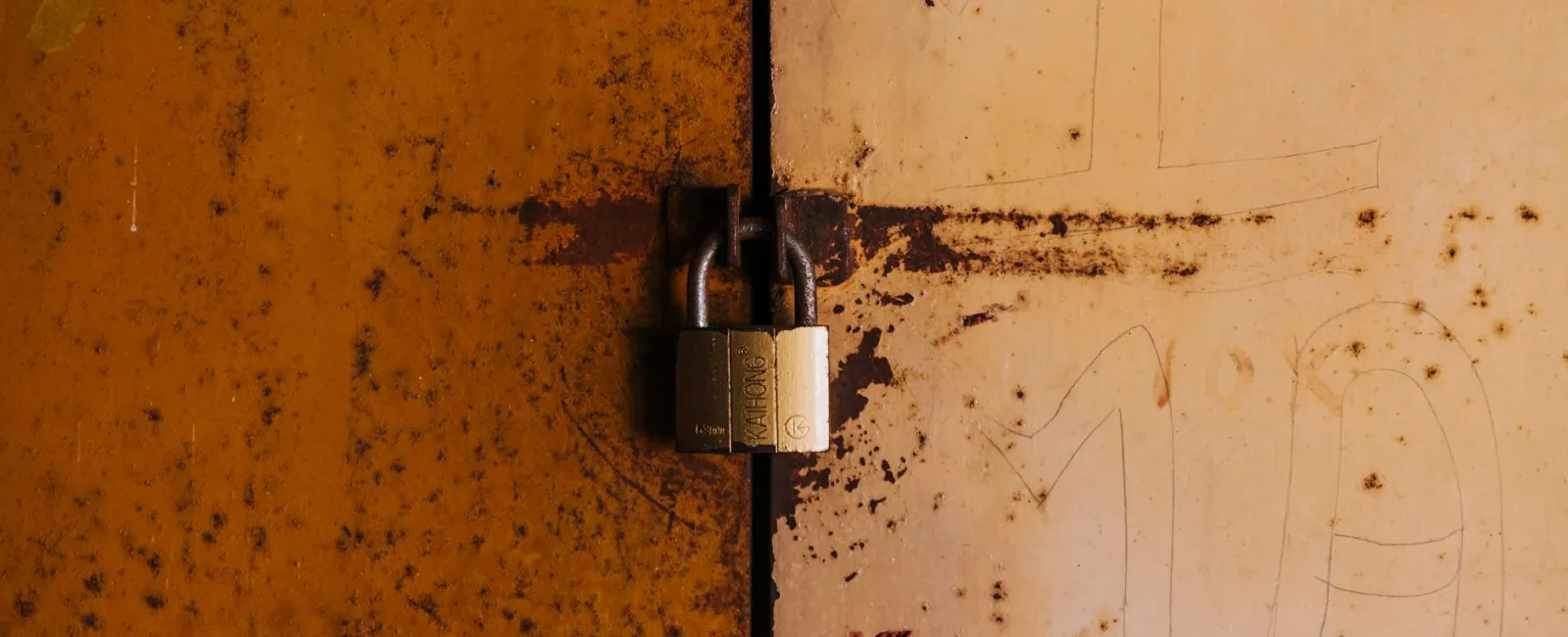 Rusty metal doors secured with a brass padlock in the center with weathered orange and beige paint.
