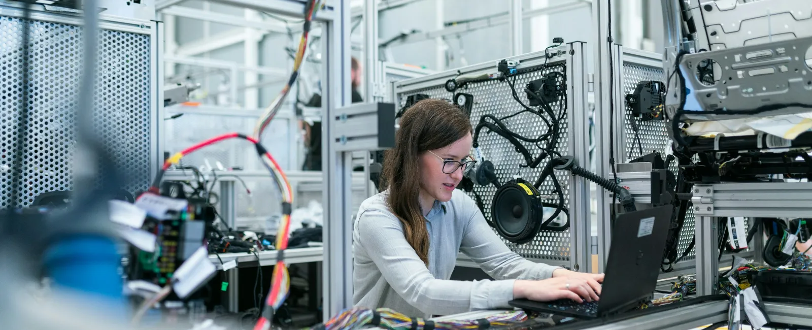 Female engineer working on laptop in a modern automotive electronics lab with circuit boards and wiring.