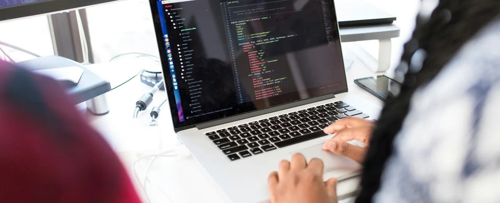 Person typing code on a laptop in a modern workspace with a monitor showing mountain scenery