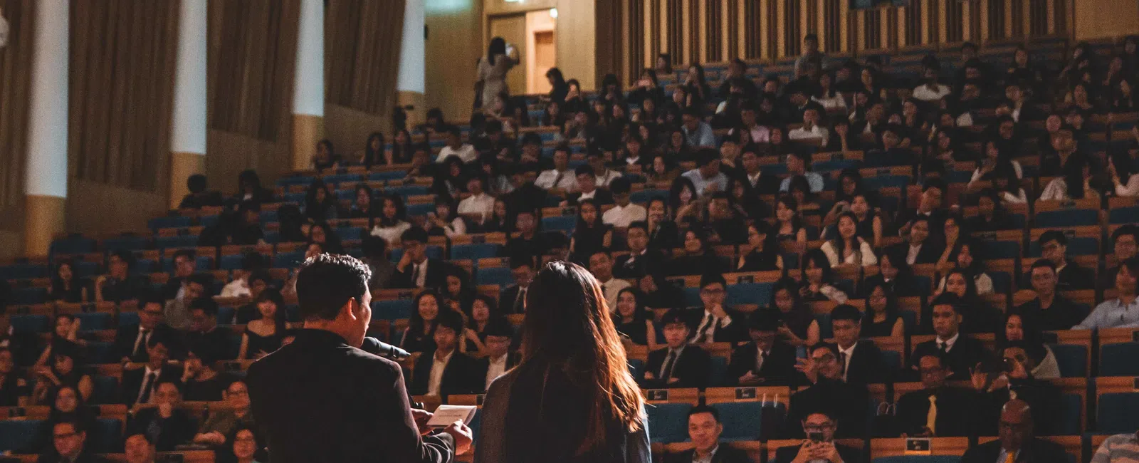 Two speakers presenting to a large audience in a dimly lit auditorium with wooden paneling and blue seats.