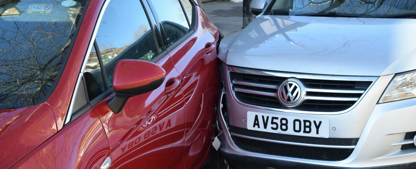 Red and silver cars involved in a close side-swipe collision on an urban street with visible damage.