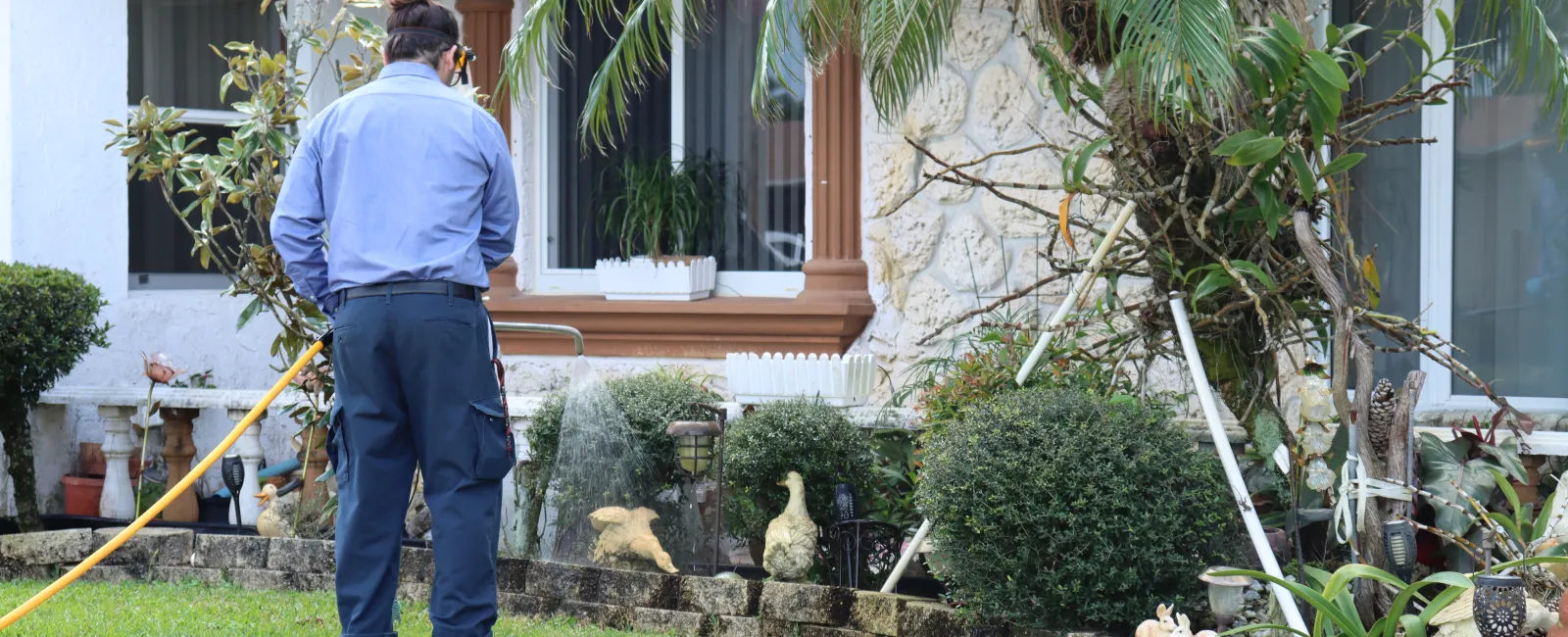 Gardener watering a residential garden with plants, shrubs, and decorative statues near a stone wall.