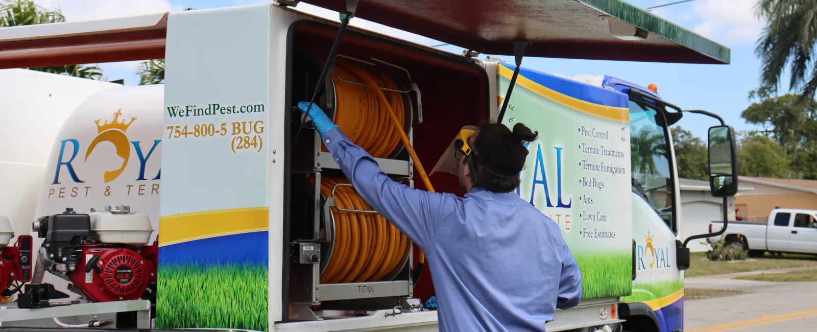 Pest control worker retrieving hose from Royal Pest & Termite truck under clear blue sky on suburban street.
