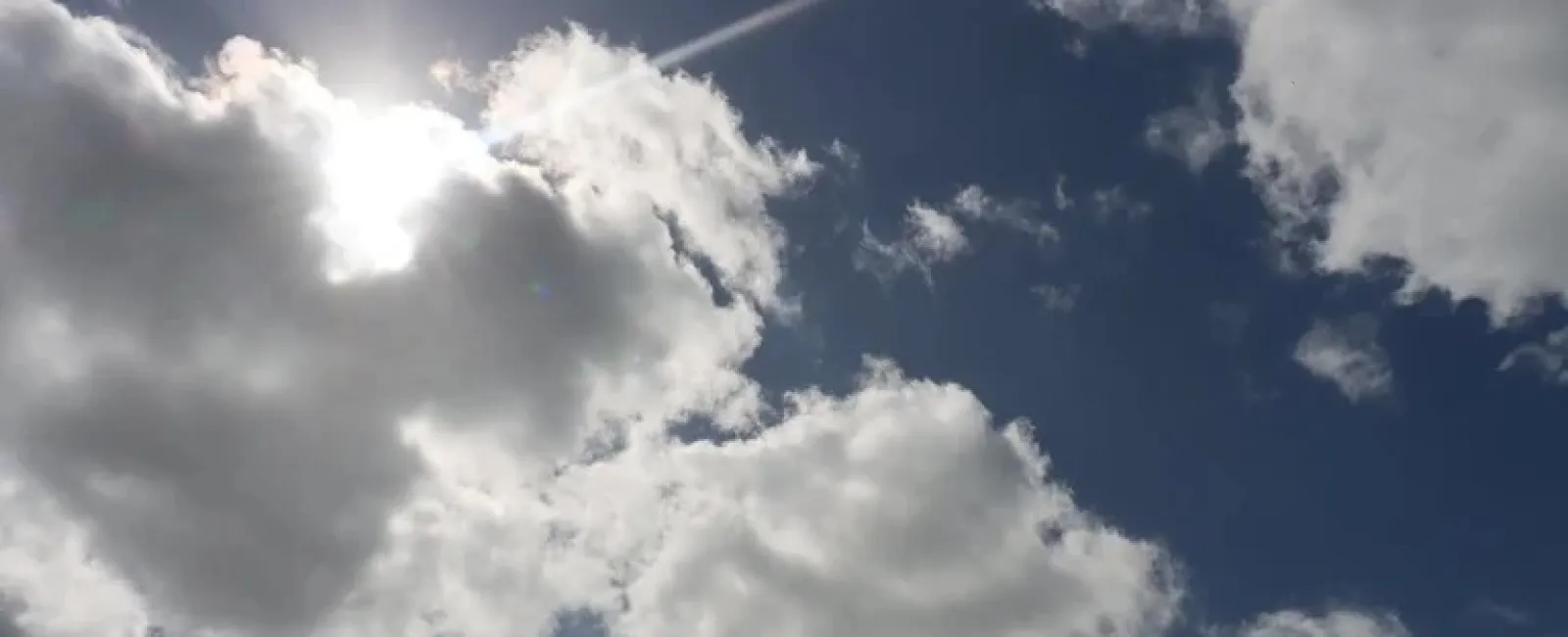 Sunlight shining behind fluffy white clouds against a deep blue sky with scattered smaller clouds.