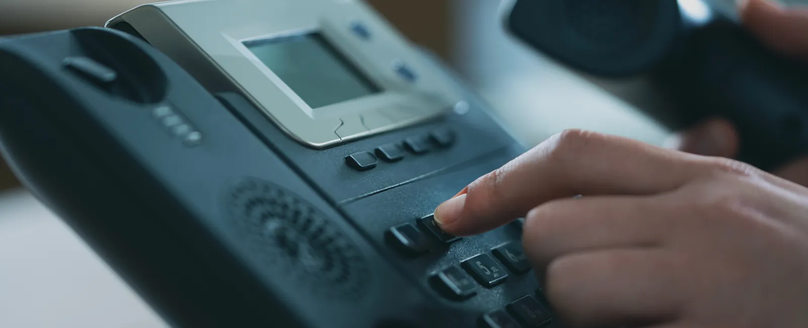 Close-up of a hand dialing a number on a black office telephone with a digital display.