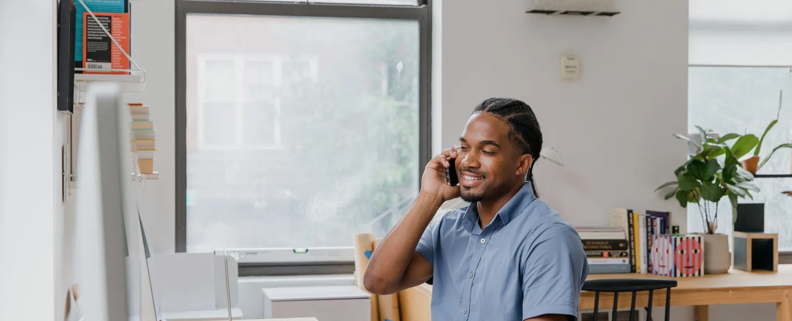 Man in blue shirt talking on phone while working at a bright home office desk with laptop and papers.