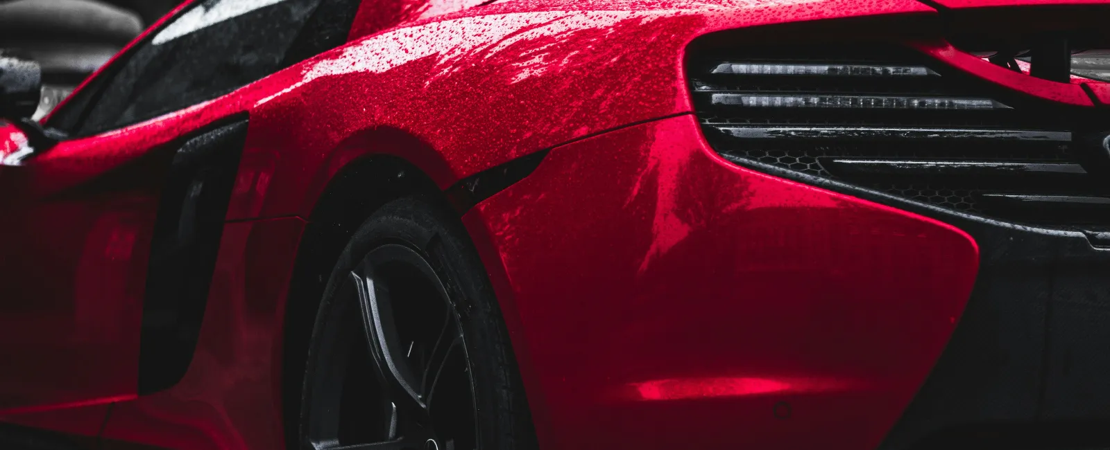 Close-up of a shiny red sports car rear with rain droplets and sleek tail light design under natural lighting.